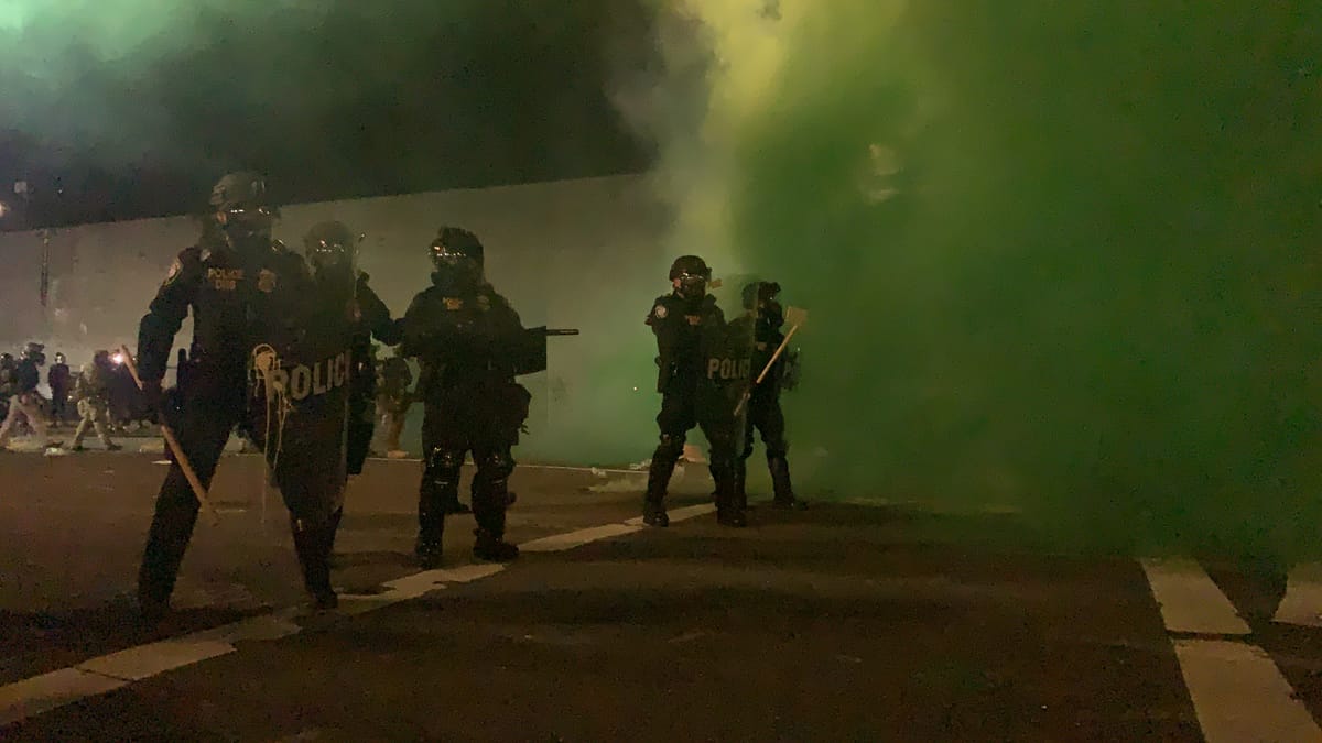 Photo of DHS police holding a line outside of Portland ICE. They are carrying shields, bats, and there is a vibrant thick green cloud of smoke behind them from munitions the feds deployed